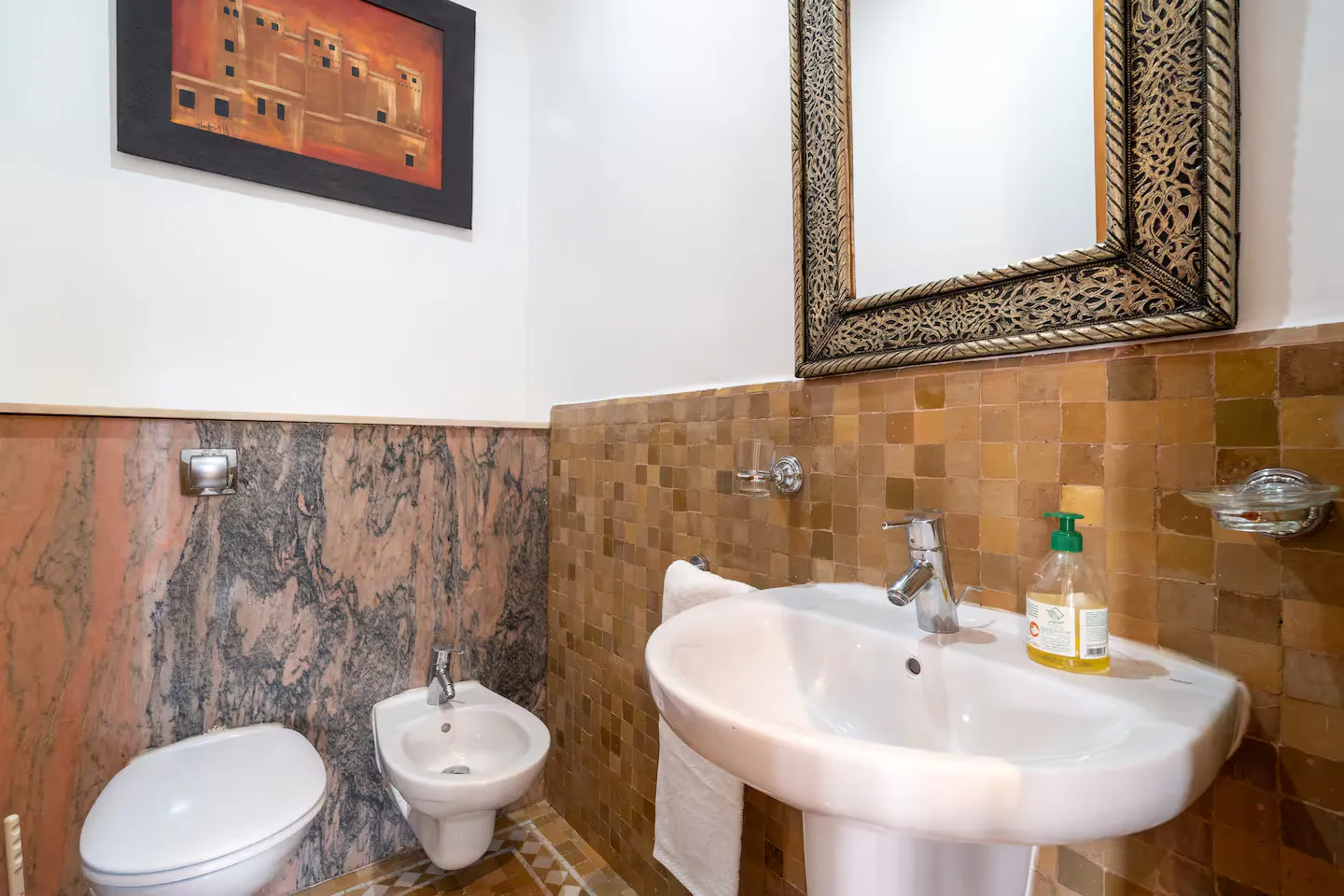 Bathroom of a vintage apartment in Marrakech with ochre mosaic tiles, a marble wall, white pedestal sink, and a traditionally framed mirror.