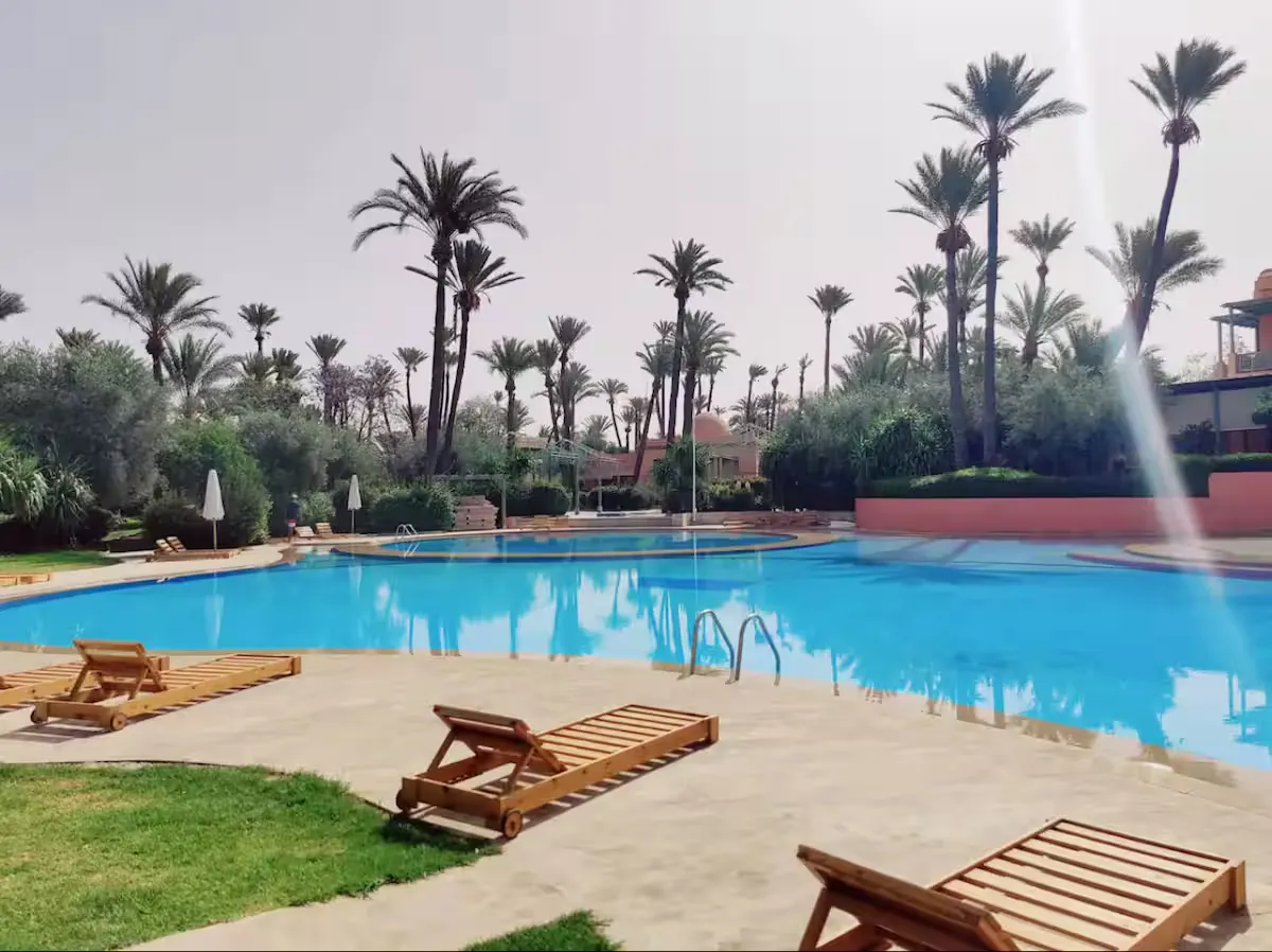 Alt : Wooden sun loungers lined up by a vast swimming pool surrounded by numerous palm trees in Marrakech, close to a vintage apartment.