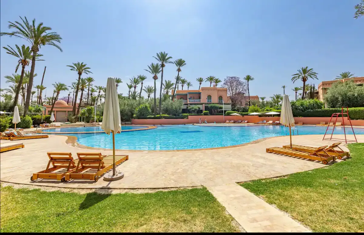 Large communal swimming pool at the Marrakech Palmeraie residence, surrounded by palm trees, with wooden sun loungers in the foreground near a vintage apartment complex