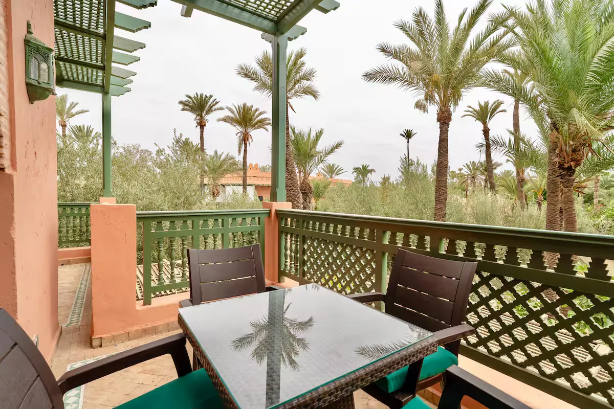Covered wooden terrace of a vintage apartment in Marrakech with glass dining table, chairs, and traditional green railing overlooking palm trees