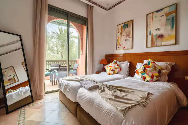 Twin bedroom in a vintage apartment in Marrakech, opening onto a terrace with palm tree views, decorated with abstract artwork and a large floor mirror.