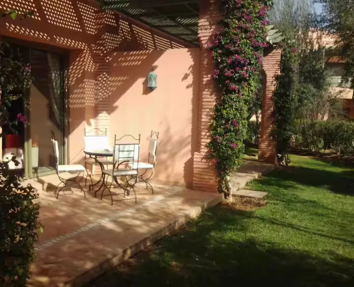 Shaded ground-floor terrace with wrought iron outdoor dining furniture opening onto the lawn of an apartment with pool in Marrakech