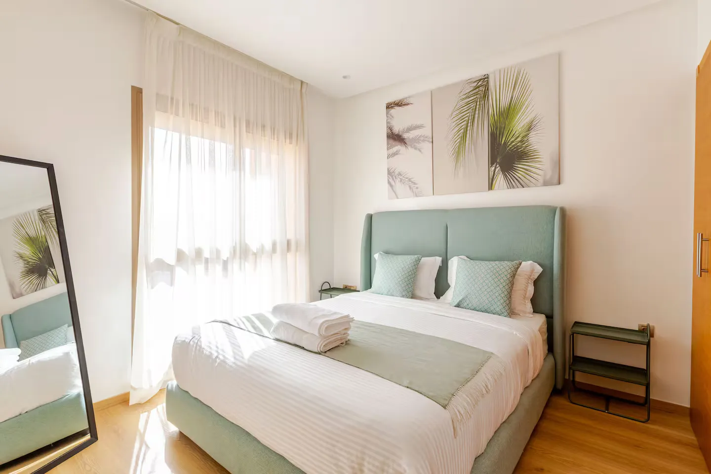 Third comfortable double bedroom with sage green headboard, full-length mirror, and tropical art in a palm grove apartment
