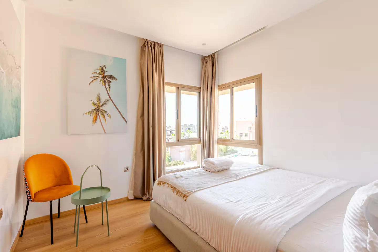 Bright double bedroom with made bed, orange velvet chair, and corner windows in a palm grove apartment