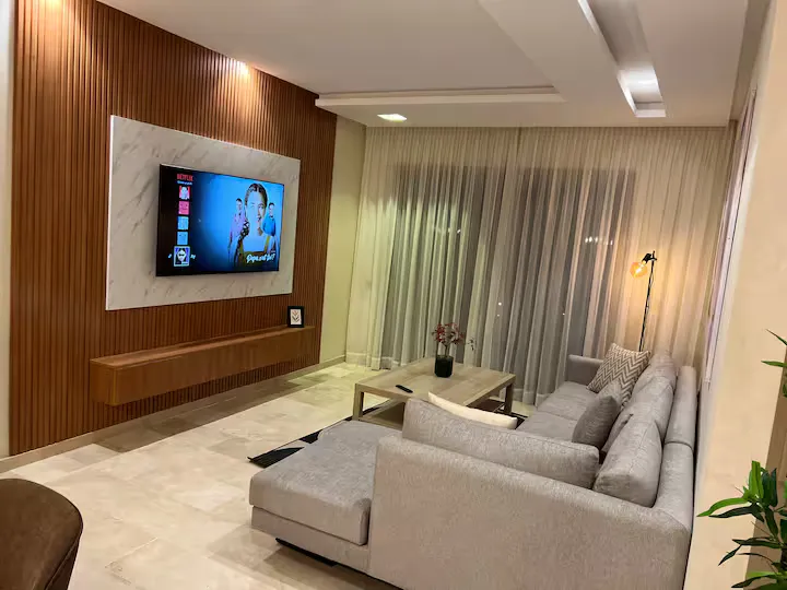 Modern living room with sectional sofa, Smart TV, and wooden wall paneling in a palm grove apartment in Marrakech